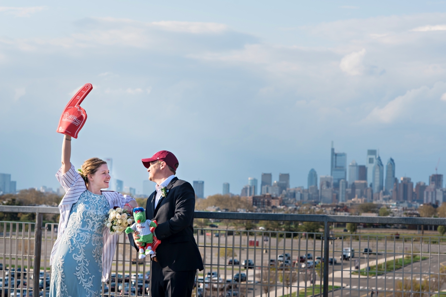 A bride and groom in front of the Philadelphia Skyline and dressed in Phillies hats and jersey.